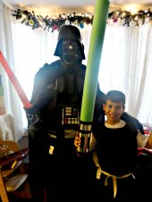 Darth vader in costume with red lightsaber in hand with Birthday boy in homemade Jedi knight robe and Star Wars pool noodle sword